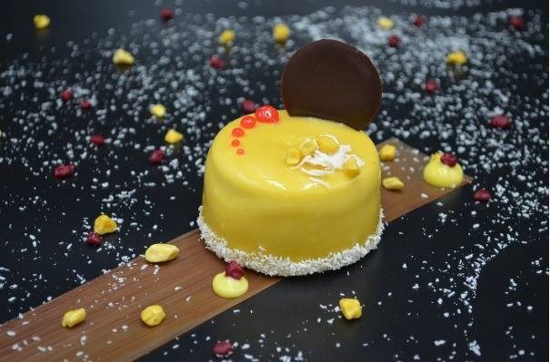 Un pan di Spagna al cacao, inzuppitura leggera al rum, mousse al cocco, cuore di mango Alphonso, glassa al mango Alphonso. Decorato con un dischetto di cioccolato fondente e scaglie di cocco disidratato